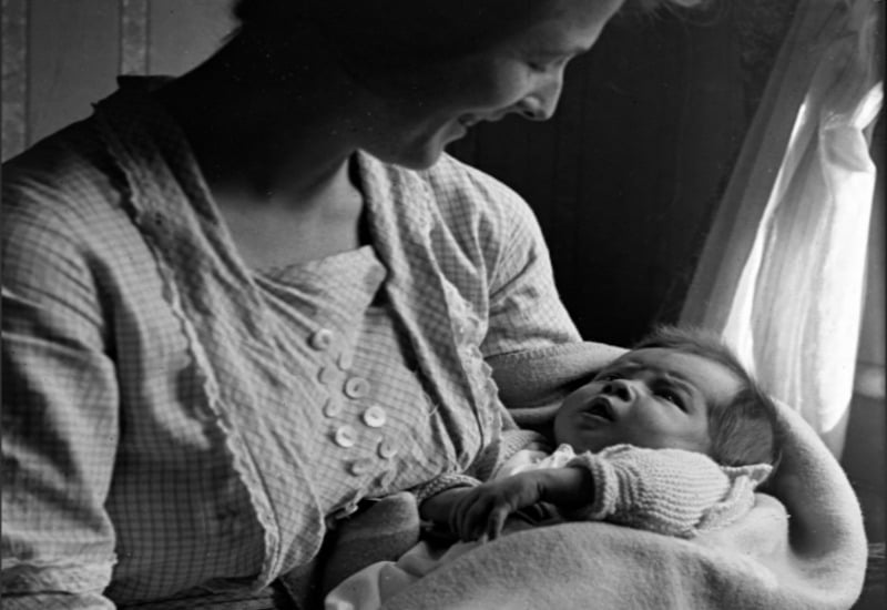 A woman smiles lovingly while holding a newborn baby wrapped in a blanket near a sunlit window. The baby looks up at her, and both appear calm and content.