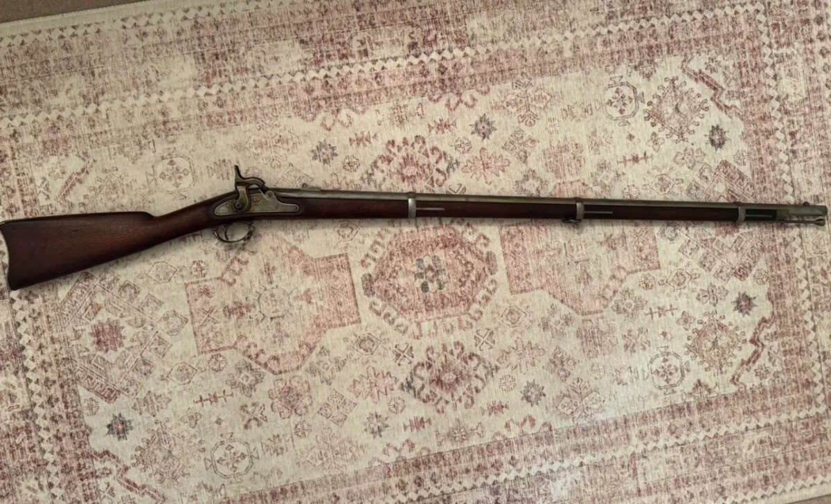 A vintage, long-barreled musket with a wooden stock and metal hardware is lying horizontally on a patterned rug with muted red and beige tones.