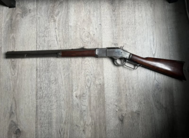 A lever-action rifle with a wooden stock and long barrel is placed on a light-colored wooden floor, viewed from above.