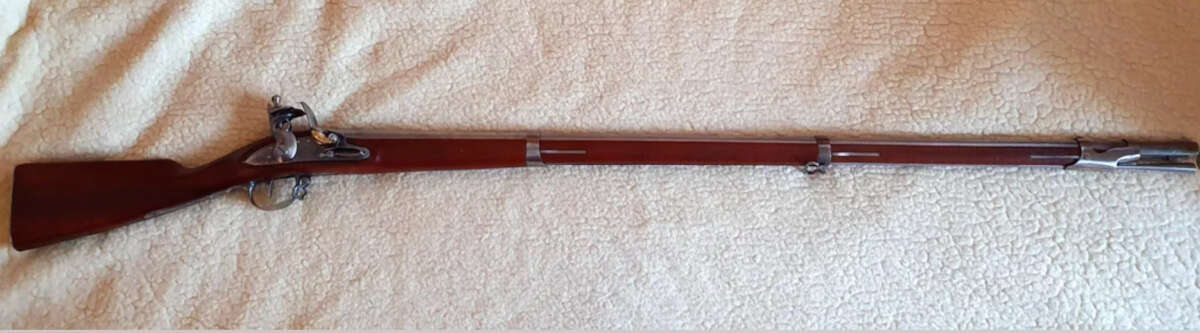 A long antique flintlock musket with a wooden stock and metal barrel lies on a light-colored textured fabric surface.