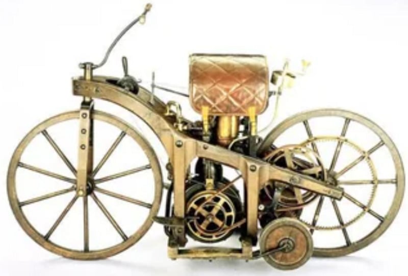 An antique, steam-powered wooden bicycle with large spoked wheels, a quilted leather seat, exposed mechanical parts, and a small rear stabilizer wheel, displayed against a white background.