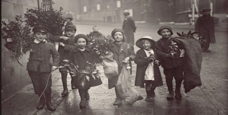 A group of six children in old-fashioned clothing smile and carry bundles of holly and greenery on a wet city street, with more people and buildings visible in the foggy background.