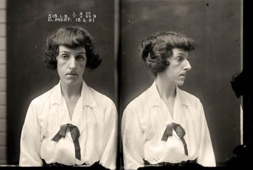 A vintage black-and-white mugshot shows a woman with short, wavy dark hair wearing a white blouse with a dark ribbon. The photo includes both front and side profiles, with identification details at the top.
