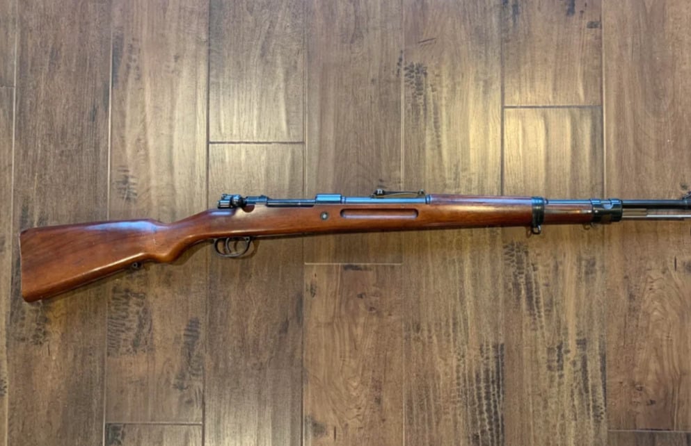 A vintage bolt-action rifle with a wooden stock and metal barrel is lying on a brown wooden floor.