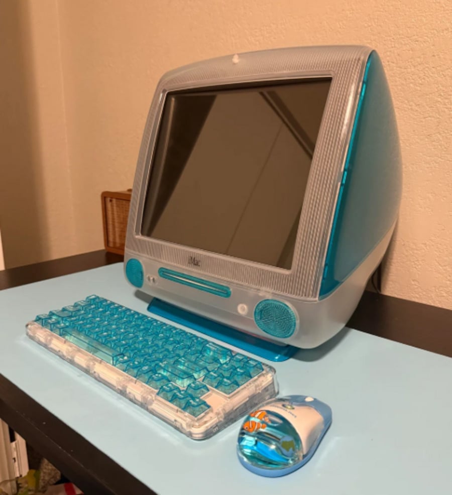 A blue and white vintage Apple iMac G3 computer sits on a desk, accompanied by a matching translucent blue keyboard and mouse.