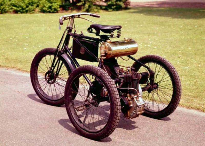 A vintage three-wheeled motorcycle with large spoked wheels, a black frame, exposed engine components, and a brass fuel tank, parked on a paved path next to grass.