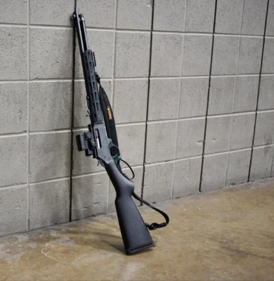 A black shotgun with a sling is propped upright against a gray concrete block wall on a polished floor inside a building.