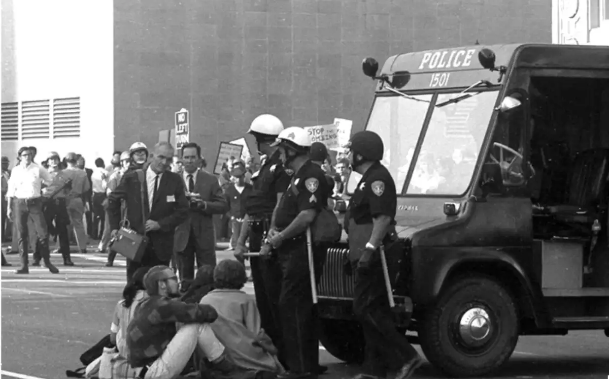 Black-and-white photo of police officers standing near a police vehicle as several seated protesters face them. A group of people and reporters with cameras observe the scene in the background. Signs are visible among the crowd.