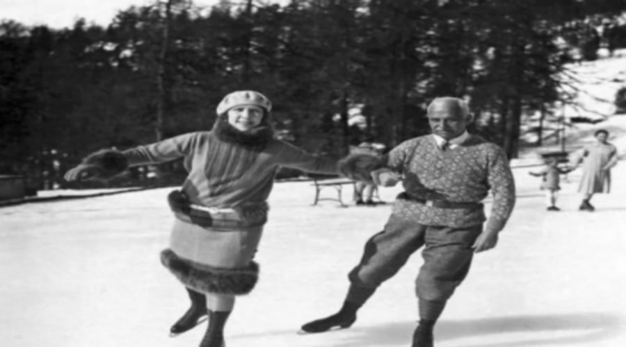 A smiling woman and man ice skate outdoors in winter clothing, holding hands. Snow covers the ground, and pine trees fill the background. Other skaters appear behind them. The photo is black and white.