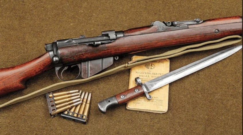 A vintage bolt-action rifle with a wooden stock is displayed with a fixed bayonet, bullets, and a manual on a brown fabric surface. A canvas sling is attached to the rifle.