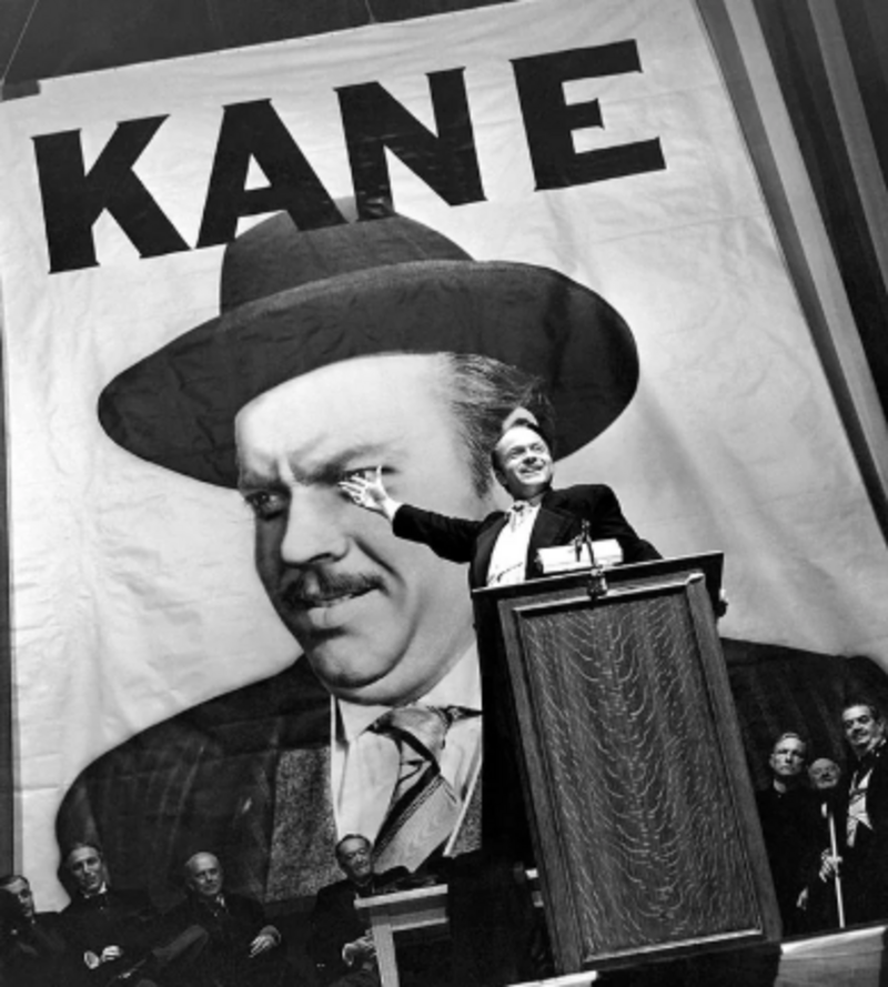 A man stands at a podium, gesturing energetically, in front of a large banner with the word "KANE" and a portrait of a man in a hat. Several seated people are visible in the background.