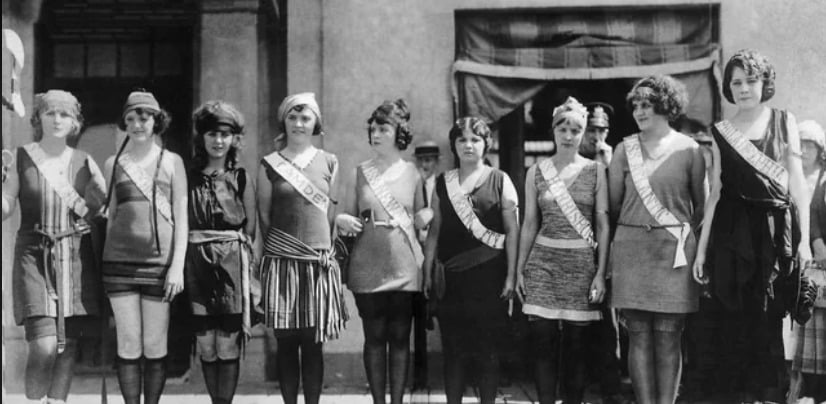 A group of women in 1920s-style dresses and sashes stand in a line outdoors, posing for a photo in front of a building with a draped window.