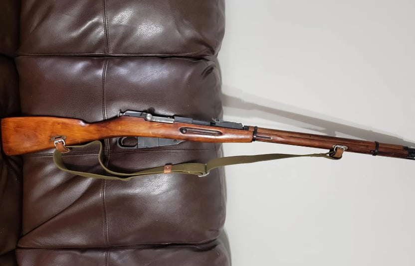 A vintage bolt-action rifle with a wooden stock and an olive green sling is resting on a brown leather couch.