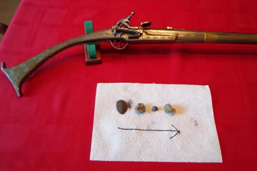 A flintlock musket rests on a red tablecloth above a paper towel displaying musket balls and metal fragments arranged in a row with an arrow pointing right.