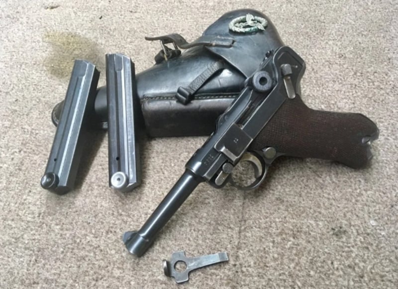 A disassembled vintage Luger pistol with two magazines, a takedown tool, and a black leather holster displayed on a beige carpeted surface.