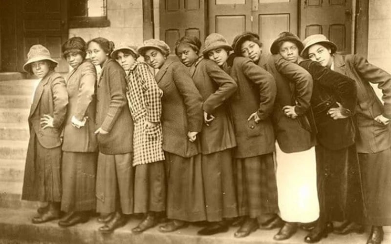 A vintage photo of nine women standing in a row, shoulder to shoulder, on steps in front of a building. They wear long coats, hats, and skirts, posing playfully and leaning slightly to one side.