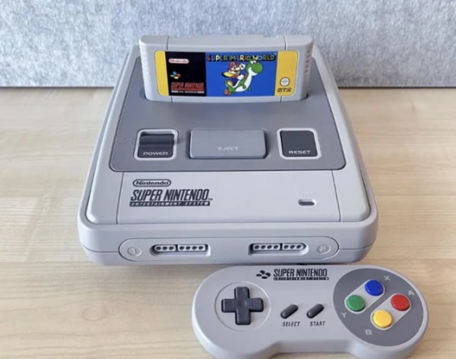 A Super Nintendo Entertainment System (SNES) console with a Super Mario World game cartridge inserted, and a SNES controller with colorful buttons placed in front of the console on a light wood surface.
