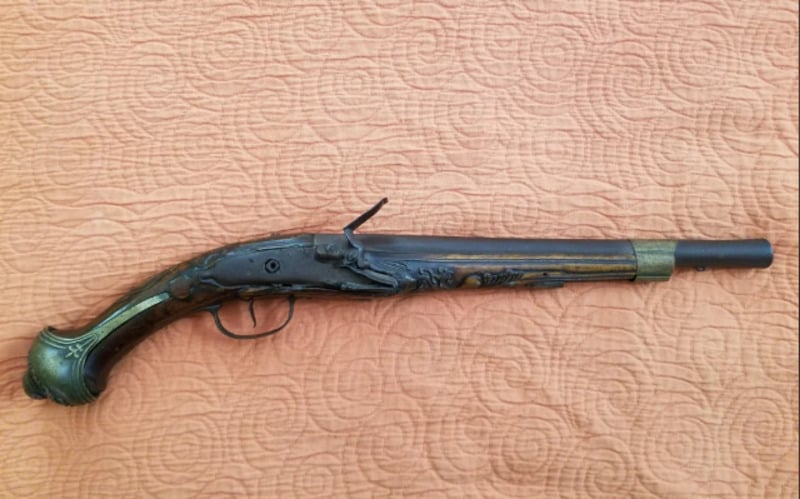 An antique flintlock pistol with ornate detailing on the handle and barrel, lying on a textured peach-colored fabric.