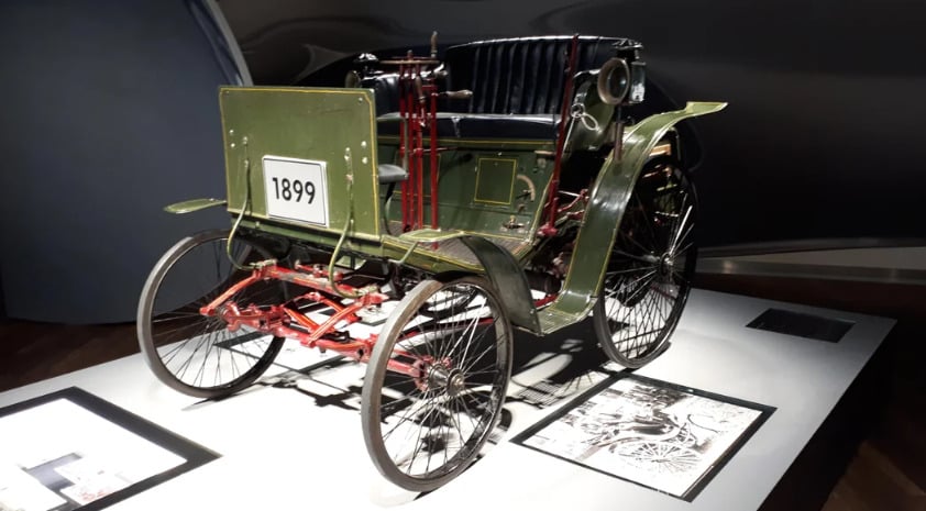 A vintage green car from 1899 with thin black tires, red undercarriage, and black leather seats is displayed in a museum with informational plaques nearby.