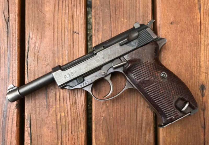 A vintage semi-automatic pistol with a dark brown textured grip and metal frame is lying on a wooden surface. The pistol appears to be worn and has markings on the slide and frame.