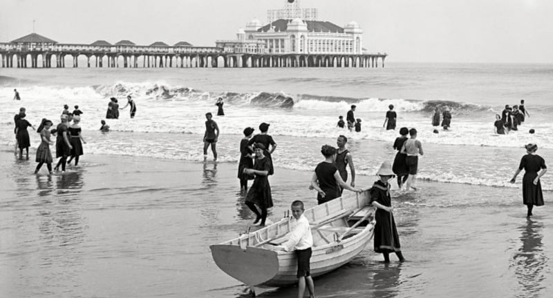 Black and white photo of people in old-fashioned swimwear wading and walking on a beach near a large pier with a grand building in the background. A boy stands beside a rowboat in the foreground.