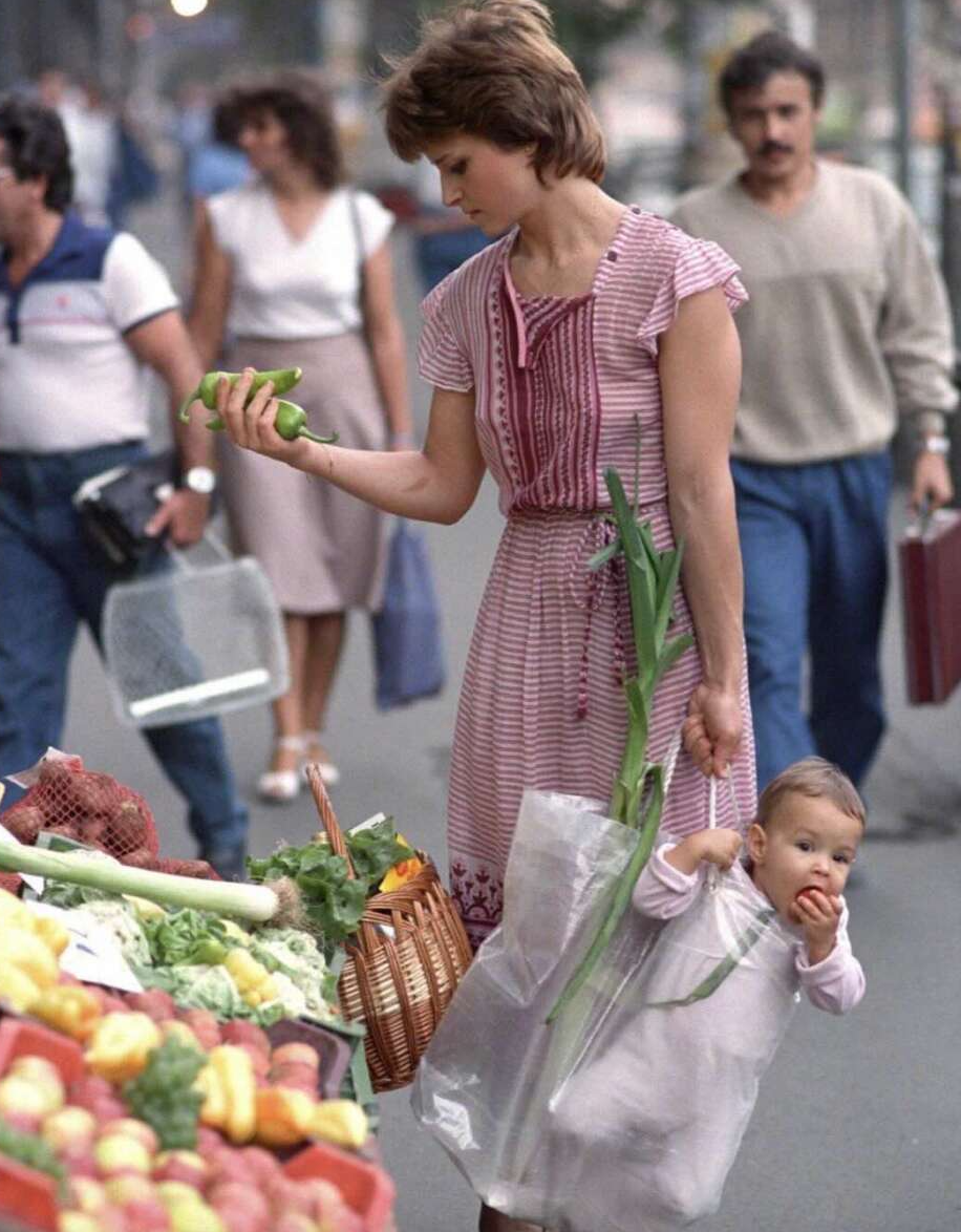 A woman in a pink dress shops for vegetables at an outdoor market, holding produce in one hand and carrying a baby in a clear bag slung from her arm; the baby bites into an apple. People walk by in the background.