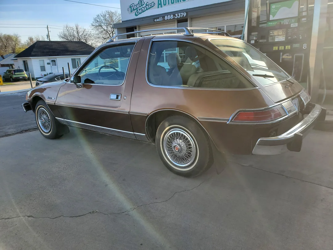 A brown classic AMC Pacer car is parked at a gas station, with a service building and other structures visible in the background on a sunny day.