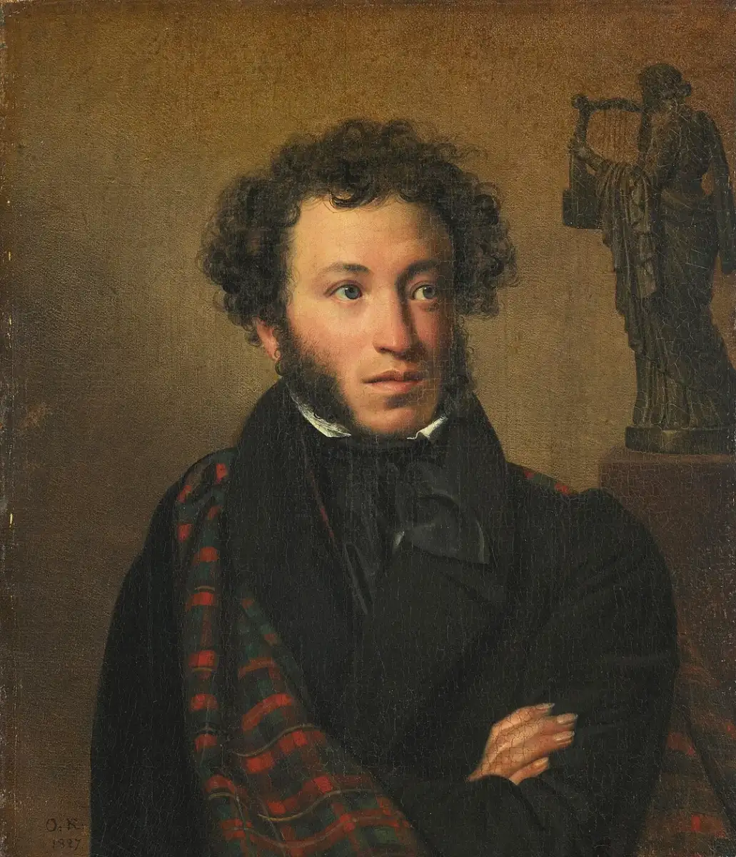 A man with curly hair and sideburns, dressed in dark clothing and a red plaid shawl, stands with arms folded in front of a statue of a woman holding a harp against a brown background.