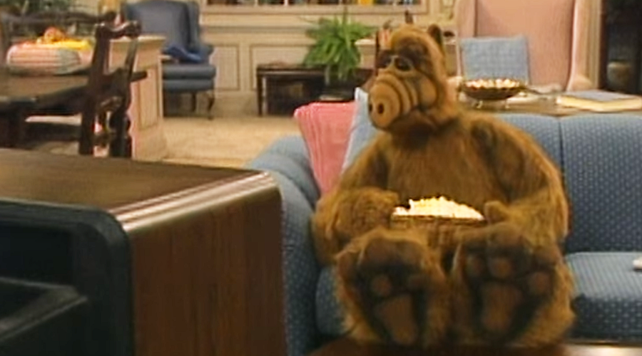 A furry, brown alien with a long snout sits on a blue couch holding a bowl of popcorn, watching TV. The setting is a cozy, lived-in living room with furniture and décor in the background.