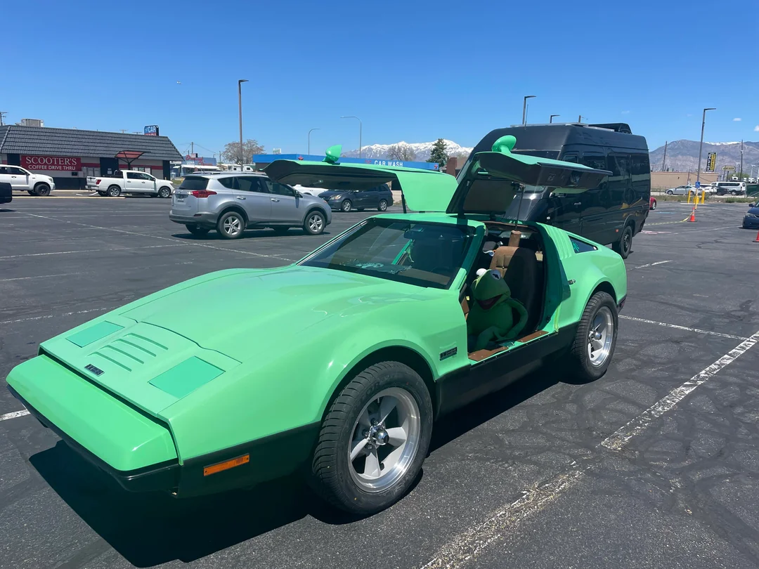 A bright green sports car with gull-wing doors open is parked in a lot on a sunny day. A person dressed as Kermit the Frog sits in the driver's seat. Other cars and shops are visible in the background.