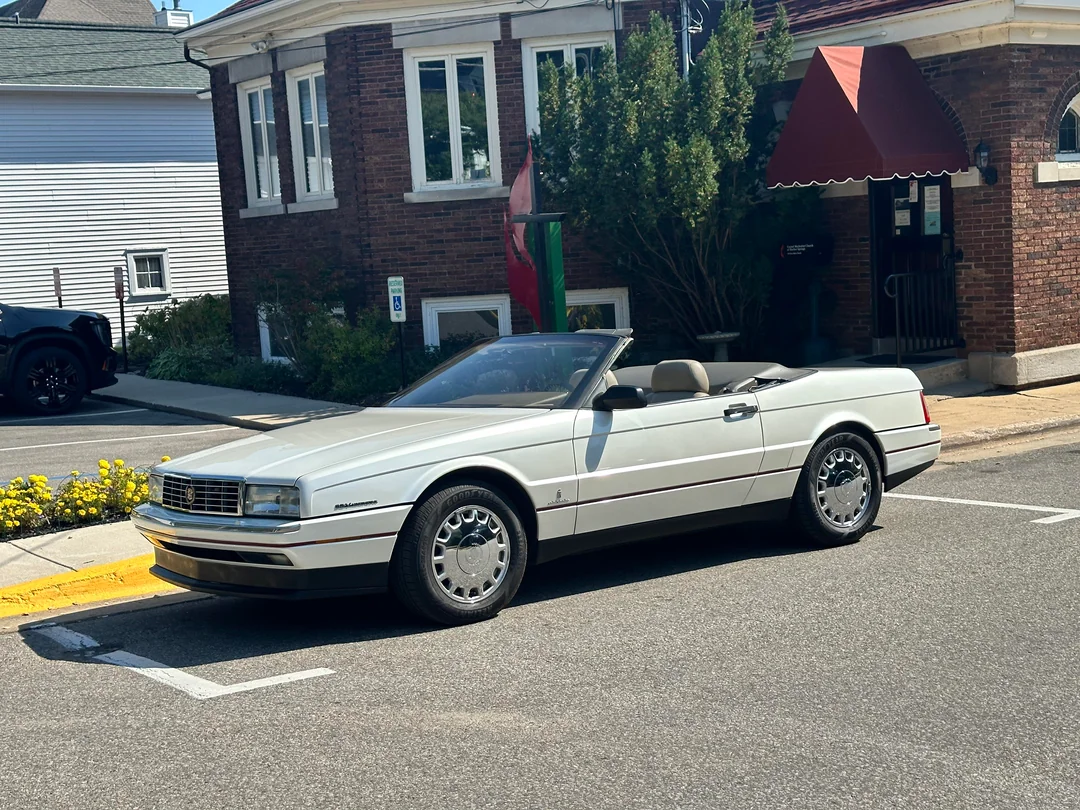 A white Cadillac Allante convertible with its top down is parked on the street in front of a brick building with a red awning and green foliage.