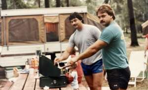 Two men with mustaches cook on a portable stove at a wooden picnic table in front of a pop-up camper in a wooded campsite. Food items and utensils are scattered on the table.