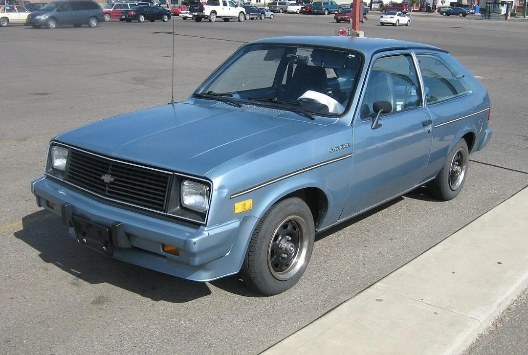 A light blue, two-door hatchback car is parked in a lot. The car has black wheels, a black front grille, and a sporty, compact design. There are other cars and a few people visible in the background.