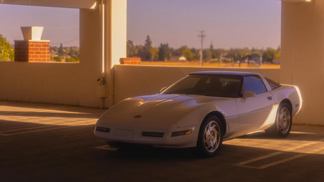 A white Chevrolet Corvette sports car is parked in a sunlit parking garage, with warm sunlight casting shadows and a suburban landscape visible in the background.