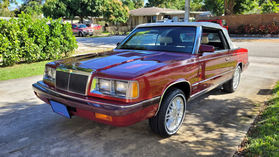 A shiny red vintage convertible car with a white soft top is parked in a residential driveway, surrounded by greenery and houses in the background under a sunny sky.