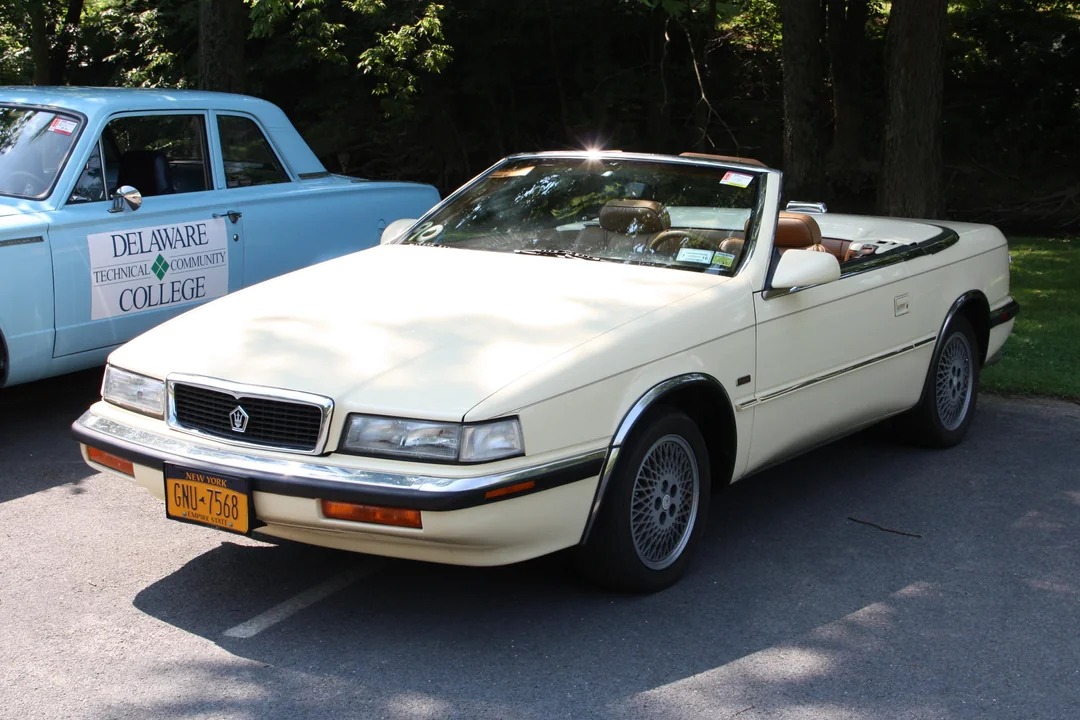 A cream-colored classic convertible car with its top down is parked on a shaded street next to a blue car. The car has a yellow New York license plate and brown interior. Trees and a sign for a college are in the background.