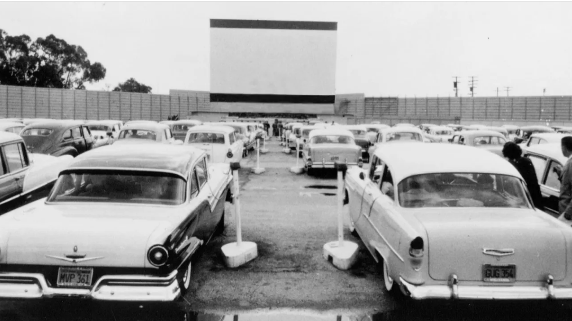 Black and white photo of a drive-in movie theater filled with vintage cars parked facing a large blank screen. People can be seen sitting in some cars, with speaker posts next to the vehicles.