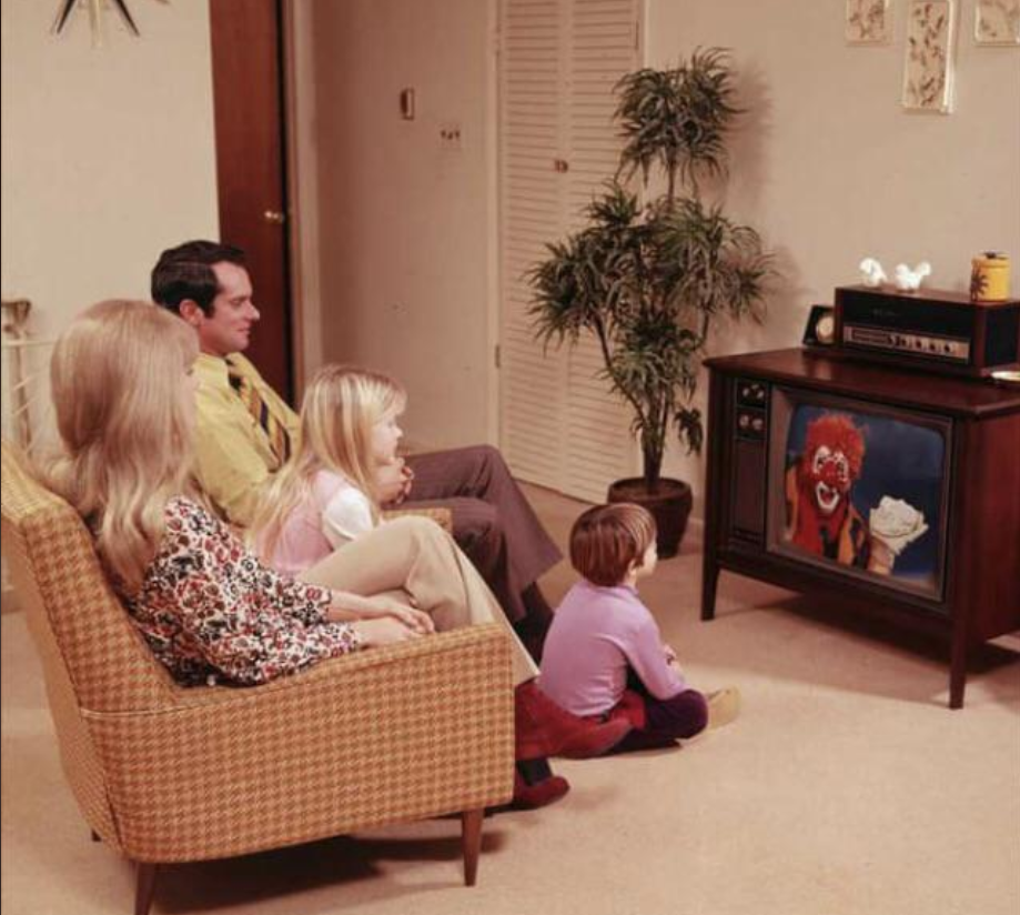 A family of four sits on a sofa and the floor, watching a TV that displays a clown with a burger and drink. The room has beige carpet, plants, and retro decor, creating a vintage atmosphere.