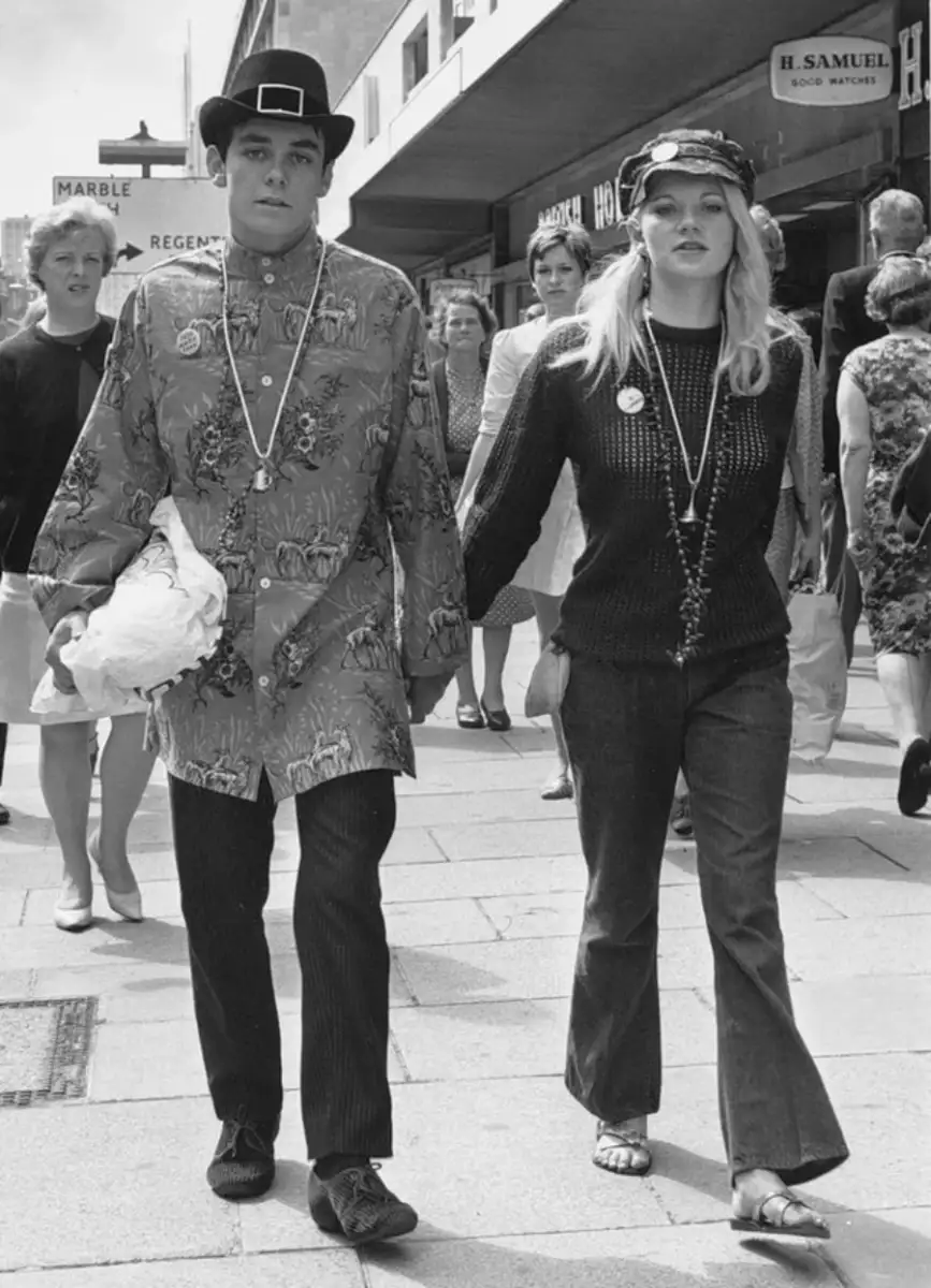 A young couple in 1960s fashion walks hand in hand on a busy street. The man wears a floral tunic and hat, while the woman wears a knit sweater, flared jeans, and a cap. People walk behind them near shopfronts.