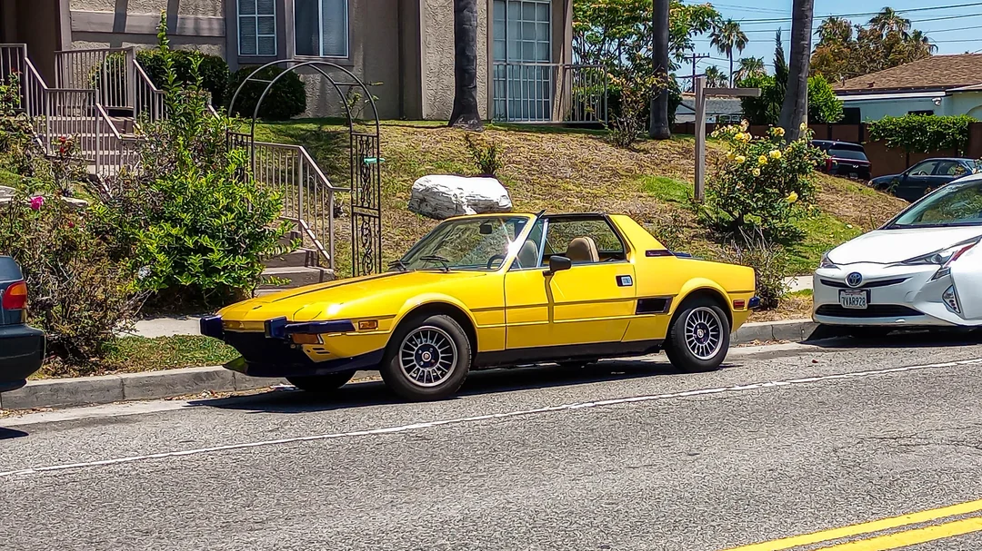 A bright yellow vintage convertible sports car is parked on a residential street between other vehicles, with houses and greenery visible in the background under a sunny sky.