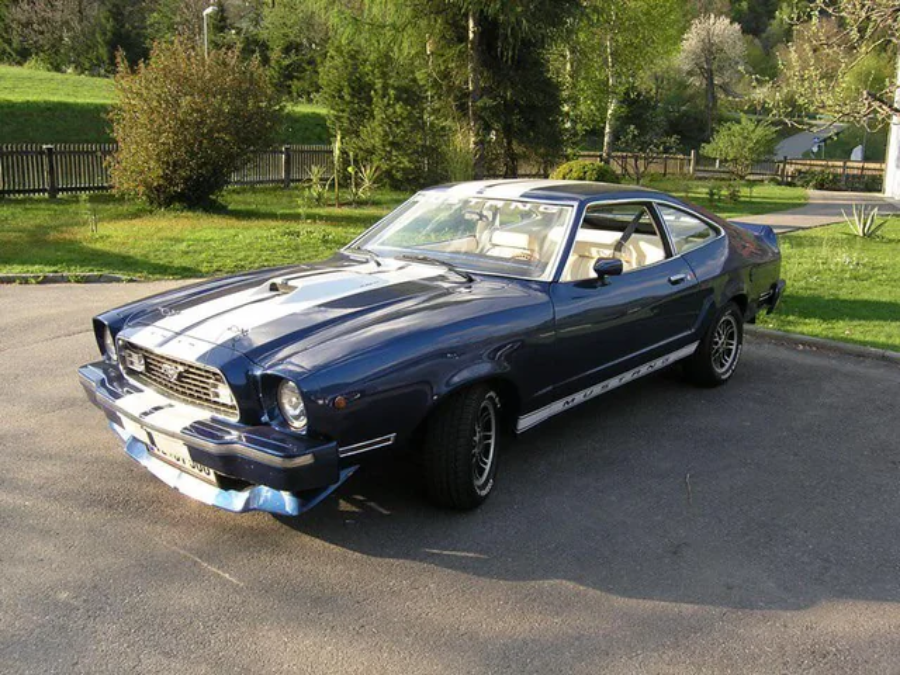 A classic dark blue Ford Mustang II is parked on a driveway near green grass and trees, with sunlight reflecting off its hood. The car has white racing stripes and vintage styling.