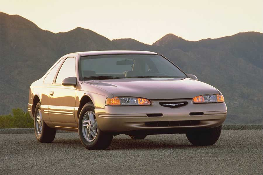 A beige two-door car is parked on a gravel road with mountains in the background at sunset.