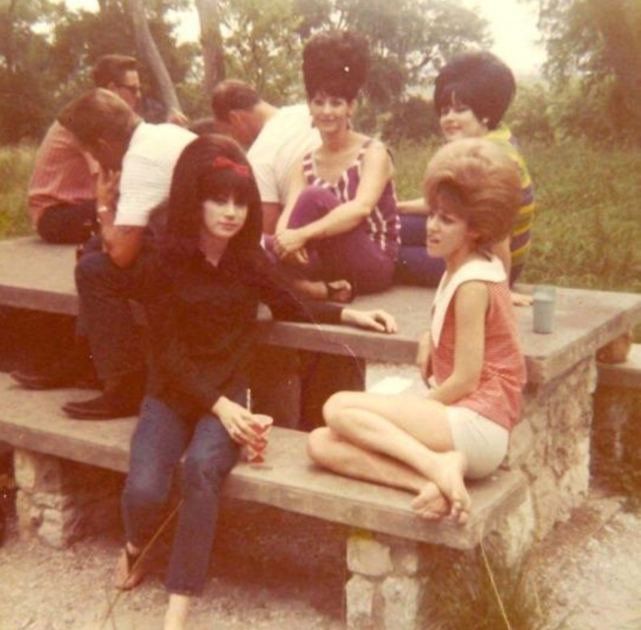 A vintage photo shows a group of people at a picnic table outdoors. Three women in the foreground wear bright clothing and have large bouffant hairstyles. Two men sit at the far end of the table with their backs turned.
