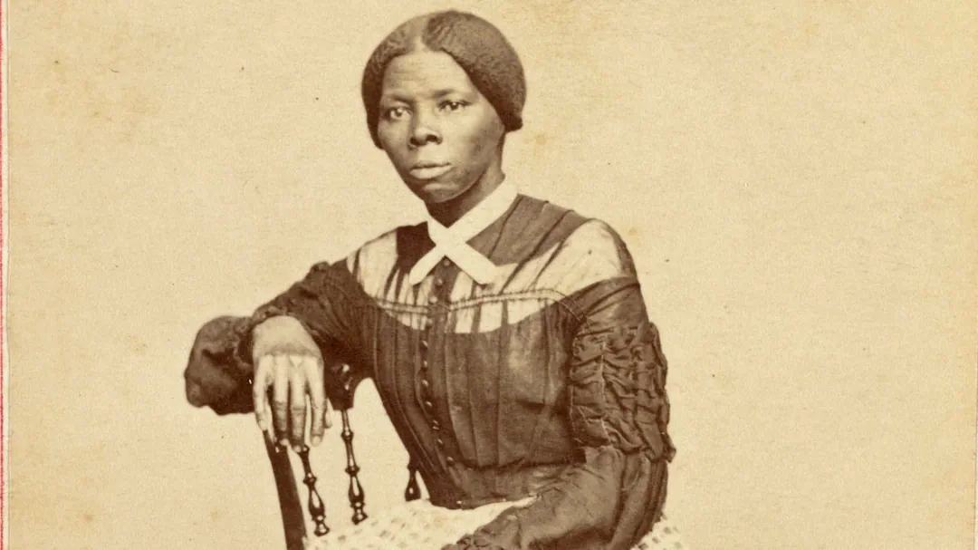 A historical sepia-toned photograph of a Black woman with her hair parted in the middle, wearing a dark dress with a white collar, seated on a wooden chair, looking solemnly at the camera.