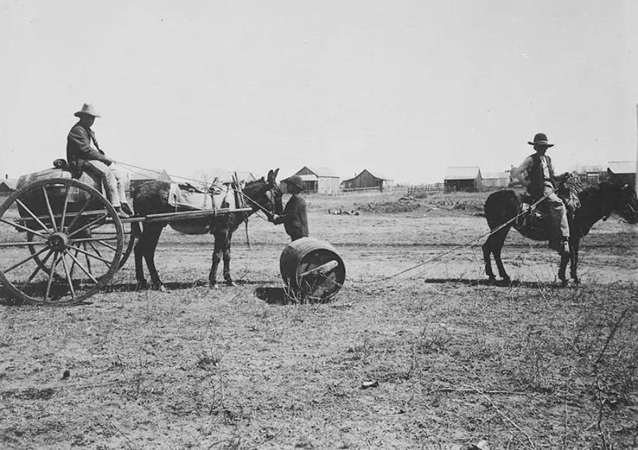 Two people wearing hats ride animals on a dirt field; one on a horse-drawn cart and the other on horseback. A barrel is dragged between them, and several small buildings appear in the background.