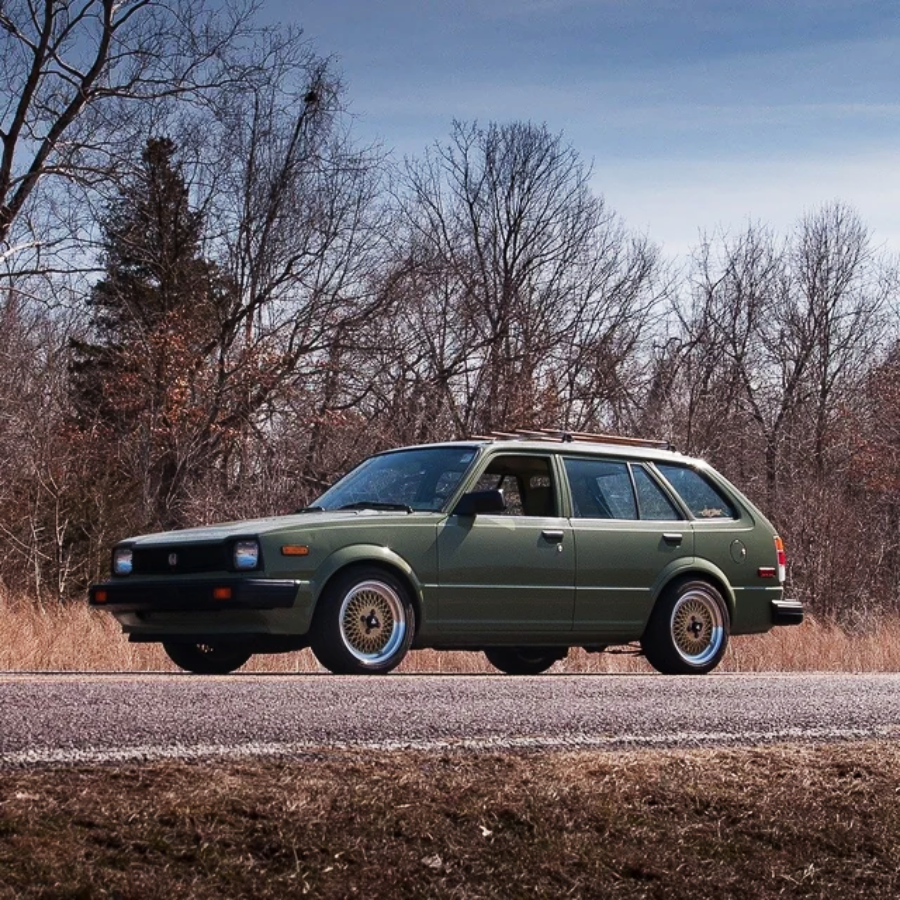 A green vintage station wagon car is parked on the roadside, with leafless trees and dry grass in the background under a partly cloudy sky.