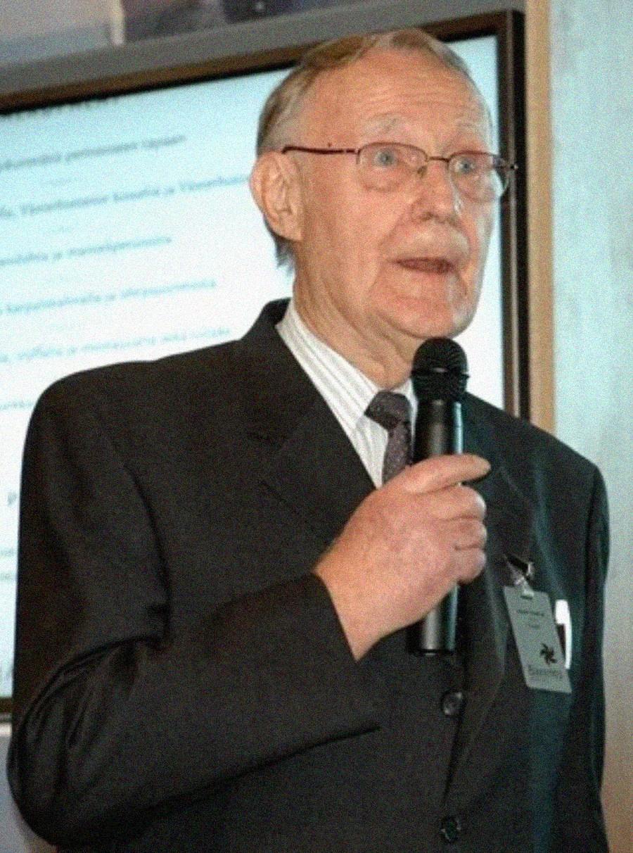 An older man in a suit and tie holds a microphone while speaking in front of a blurry presentation screen. He wears glasses and a name badge.