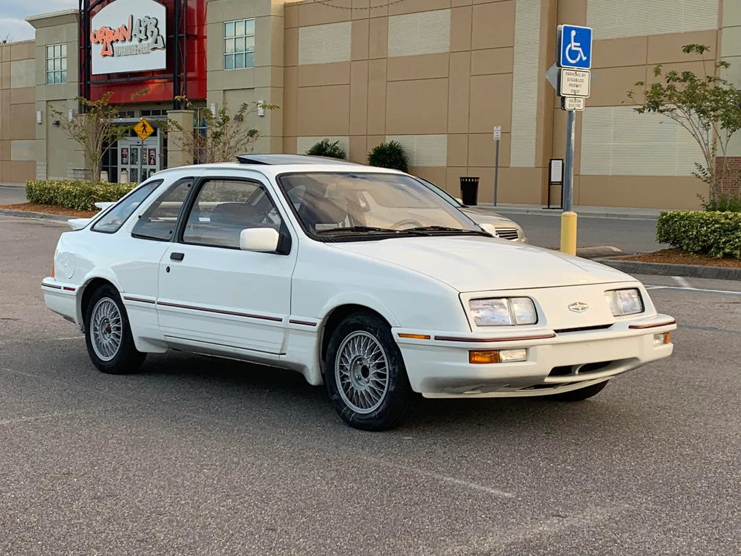 A white 1980s two-door car is parked in a lot near a shopping center. The car has pop-up headlights, alloy wheels, and a sunroof. A handicap parking sign and some trees are visible in the background.