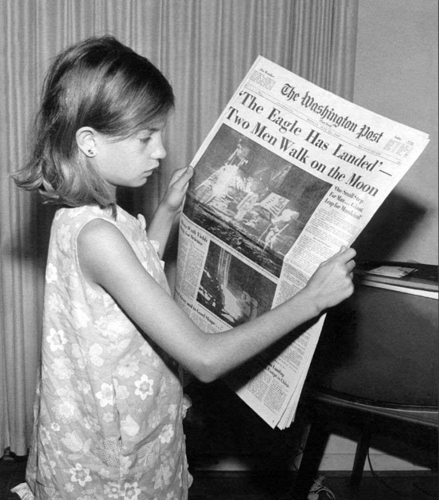 A young girl in a sleeveless floral dress reads a large newspaper with the headline "'The Eagle Has Landed!' Two Men Walk on the Moon" and photos of the 1969 Apollo 11 moon landing.