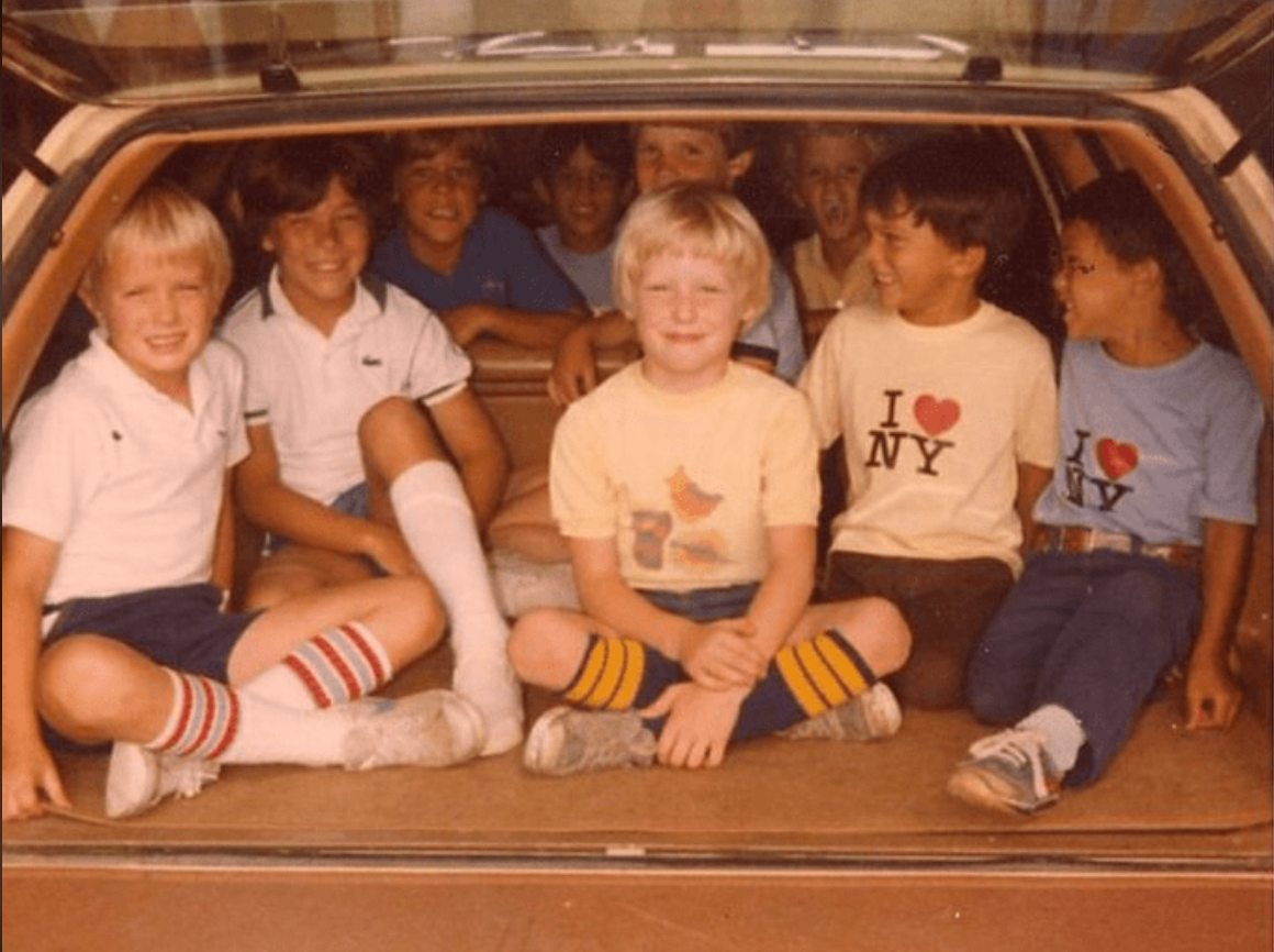 A group of eight young boys sit together in the open back of a station wagon, smiling and laughing. Some boys wear “I ♥ NY” T-shirts and striped socks, giving the image a vintage, 1970s or 1980s feel.